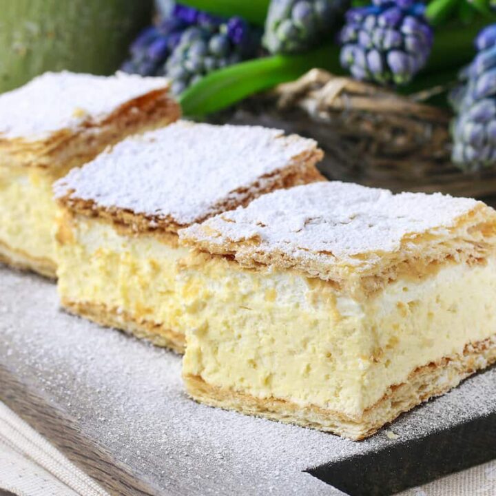 Three squares of pastry with powdered sugar on a wooden board, also known as Polish cream papal cake.
