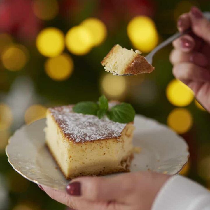 A person holding a fork with a piece of Polish cheesecake sernik over a plate, against a festive bokeh background.