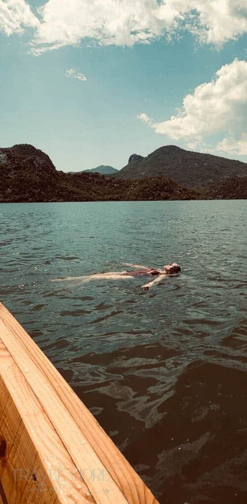 Person floating on their back in calm lake waters surrounded by green mountains under a partly cloudy sky — A peaceful outdoor experience and a relaxing option on the list of things to do in Budva.