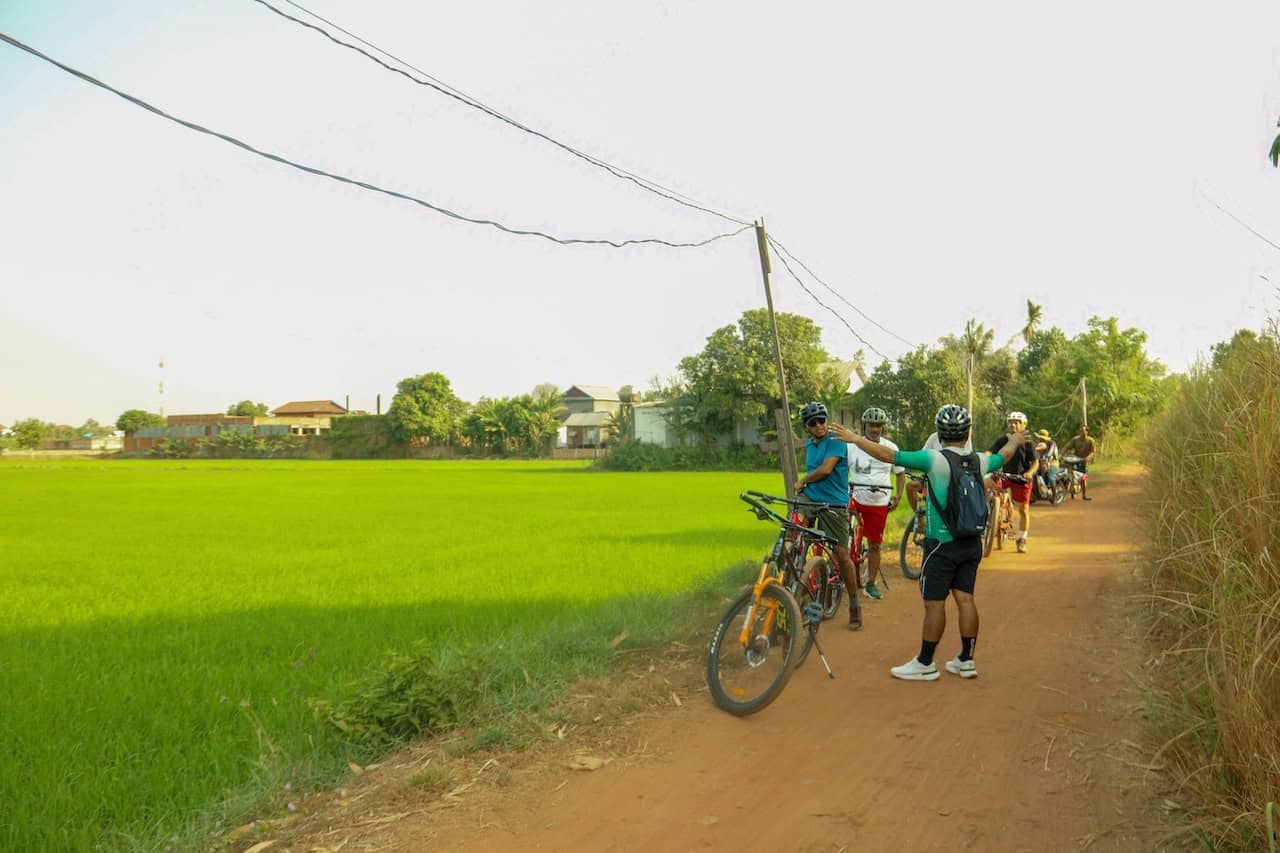 Cyclists riding along backroads between Cambodia and Laos, passing rural villages, rice paddies, Mekong River scenery, and peaceful countryside landscapes on a cross-border bike adventure.