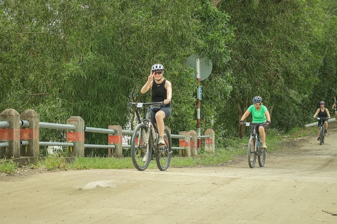 Cyclists riding through rural Cambodian villages between Siem Reap and Phnom Penh, passing rice fields, traditional Khmer houses, local markets, and countryside backroads.