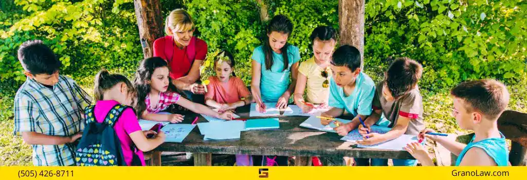 A group of children and a teacher drawing together at a picnic table outdoors in a sunny park, with the Grano Law Offices, P.C., logo and phone number.