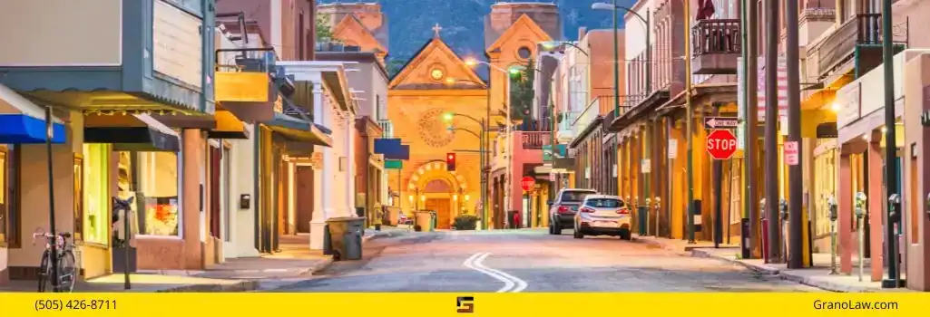 A street view at dusk in Santa Fe, New Mexico, looking down a road towards the Cathedral Basilica of St. Francis of Assisi. The street is lined with shops, cars, and a stop sign. The image footer has a yellow bar with "(505) 426-8711" on the left, a logo in the center, and "GranoLaw.com" on the right.