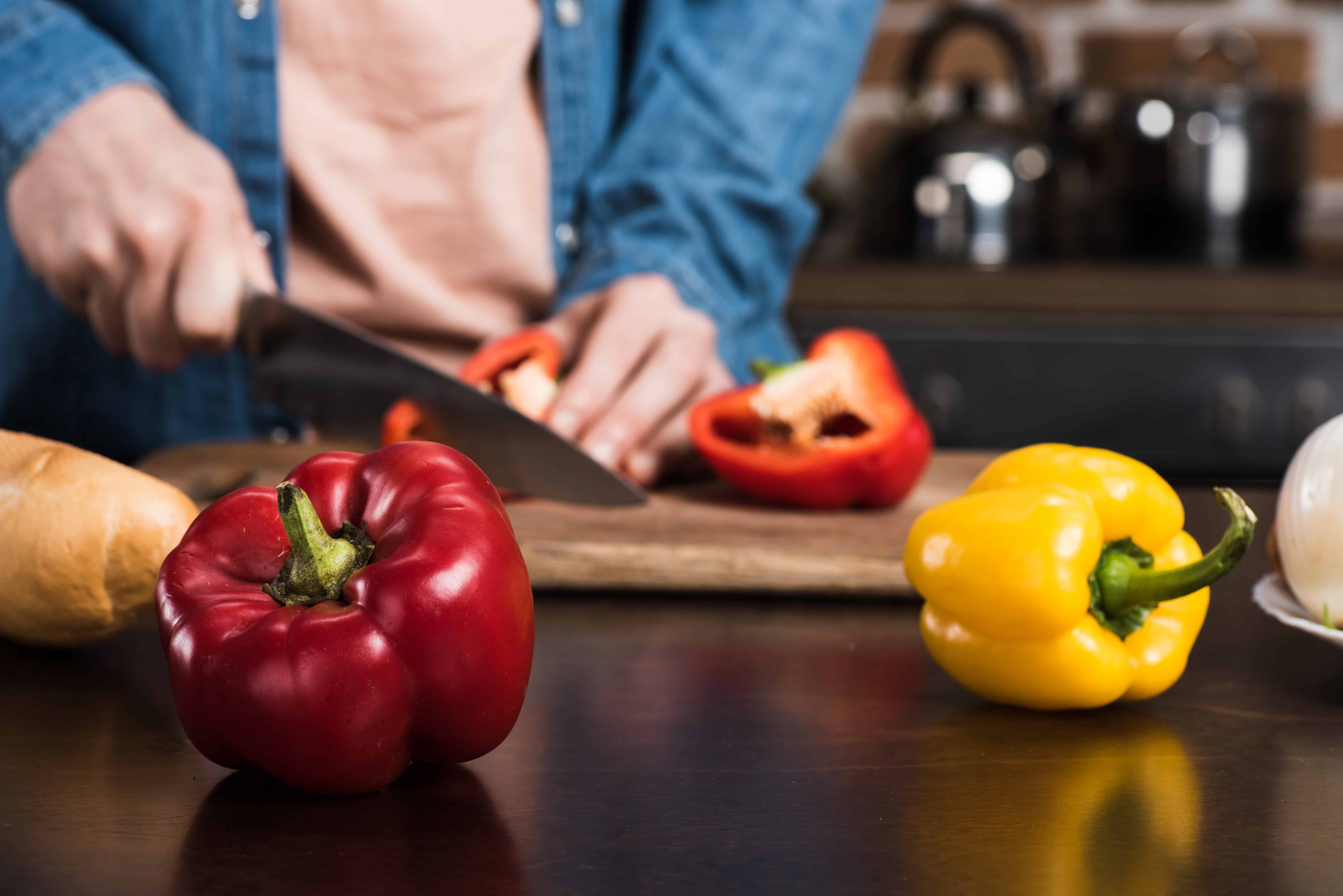man cutting a red pepper on a wooden chopping board in the background with a yellow pepper and red pepper in front closer to the camera.