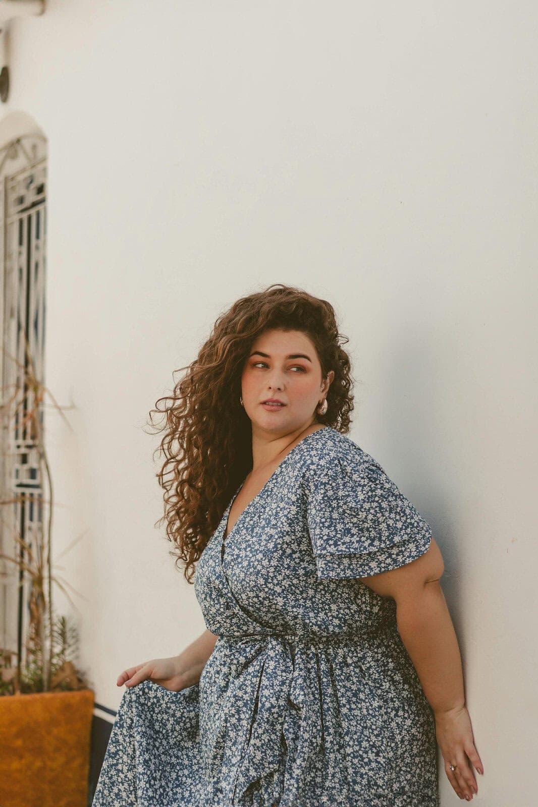 A woman with long curly hair wearing a blue floral dress leans against a white wall, looking off to the side. There is a decorative metal window and a potted plant in the background.