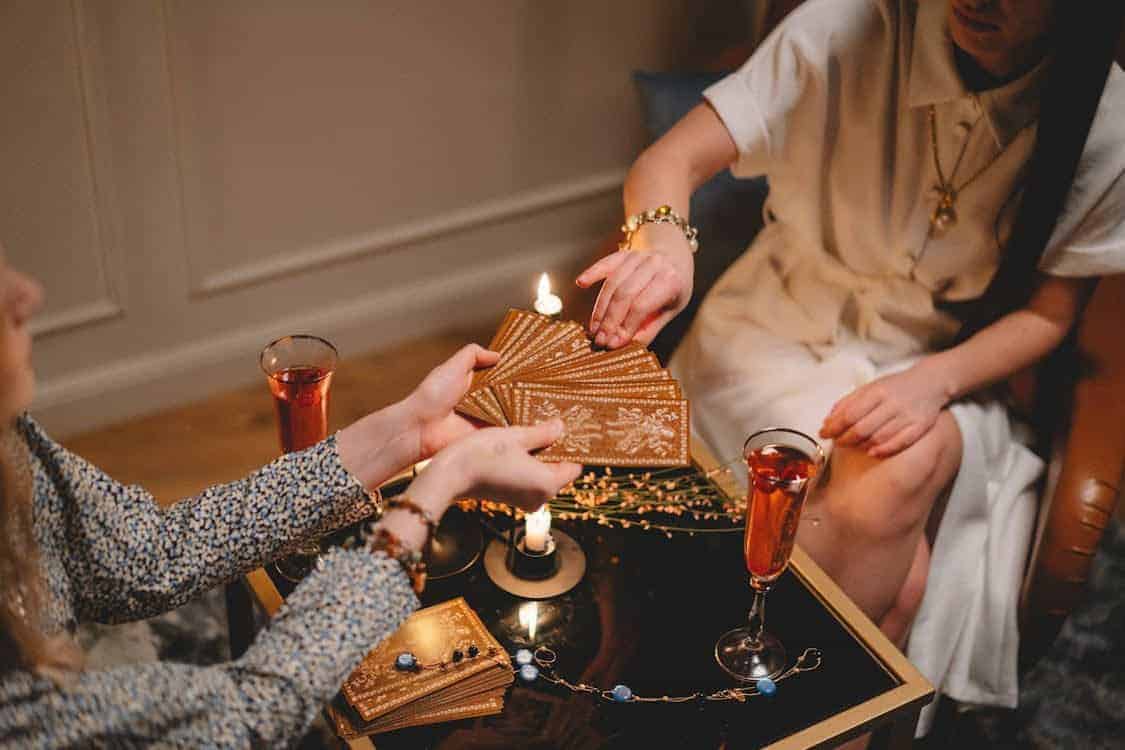 Two women sit at a table with tarot cards, lit candles, and drinks. One woman fans out the cards while the other chooses one, suggesting a fortune-telling or tarot reading session.