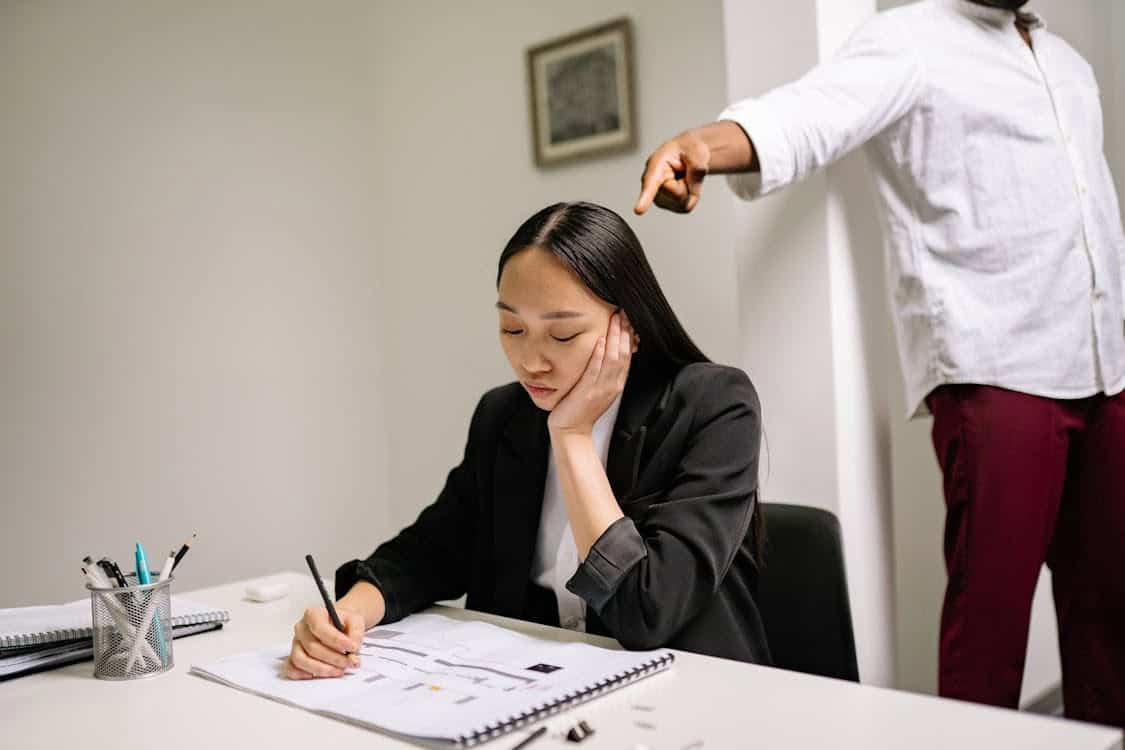 A woman in business attire sits at a desk, looking down and writing on paper with a pen, while a person standing behind her points a finger in her direction. The scene appears to be in an office setting showing workplace bullying.