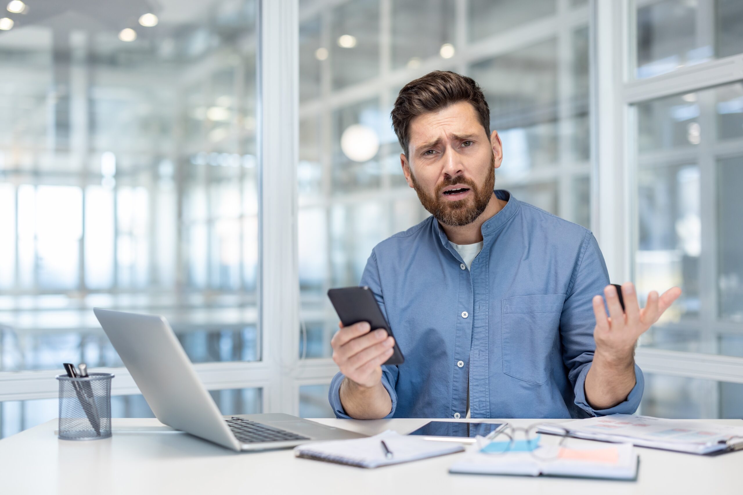 Upset and disappointed man with phone in hands. office worker at workplace looking anxiously at camera, working with laptop inside office at workplace