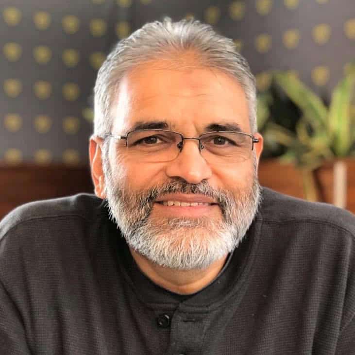 Afzul Soobedaar, founder and director of Perispec Consulting, smiling confidently in a grey polo with glasses and grey hair and beard, seated in a warm, professional setting. Afzul is an experienced strategy consultant and workshop facilitator based in South Africa.
